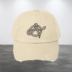 Könnte beinhalten: Beige Baseball Cap mit abgenutztem Schirm. Die Kappe zeigt eine schwarze Grafik eines stilisierten Buchstabens "A", der mit einer Schlange verschlungen ist. Die Kappe hat eine neutrale Farbe und ein klassisches sechsteiliges Design.