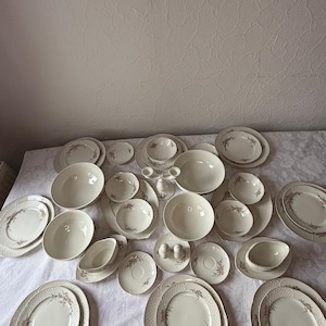 Könnte beinhalten: Eine Sammlung weißer Porzellan-Essgeschirr mit floralem Design. Das Set umfasst verschiedene Teller, Schalen, Untertassen und Servierschalen. Die Ränder der Teller haben ein strukturiertes Muster.