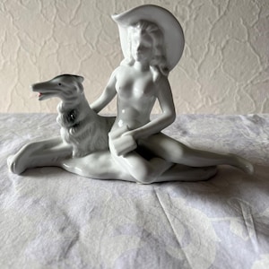May include: A white porcelain figurine depicting a reclining figure wearing a hat, with a dog. The figurine is detailed with smooth surfaces and subtle shadows, showcasing the form and texture of the sculpture. The dog has a red tongue.