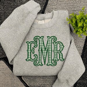 May include: A heather gray crewneck sweatshirt featuring a large, embroidered monogram of the letters EMR. The letters are filled with a green and white gingham check pattern and outlined in dark green thread. A small white size tag is visible.