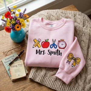 May include: A light pink sweatshirt featuring an embroidered design of a yellow pencil, a red apple, blue scissors, and a pink alarm clock above the text Mrs. Smith. The sleeve displays a yellow bow. The garment is displayed on a textured surface.