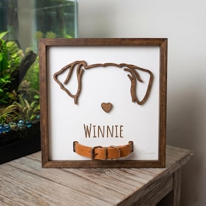May include: Wooden framed artwork featuring a dog face outline, a heart, and the name "Winnie". A brown leather dog collar is at the bottom. The frame is dark brown, and the background is white.