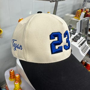 May include: A cream-colored baseball cap with a black brim, featuring the number 23 embroidered in bold blue thread on the front panel and the name Tegan embroidered in blue cursive script on the side panel of the structured crown.