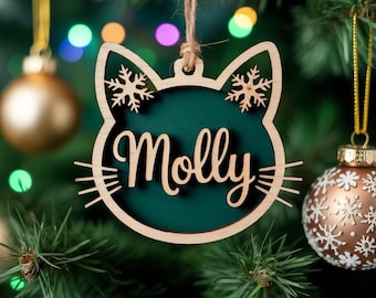 Puede incluir: Adorno navideño de madera con forma de gato con el nombre "Molly" en cursiva. El adorno tiene un fondo verde y detalles de copos de nieve. Cuelga de una cuerda de hilo en un árbol de Navidad, con bolas doradas y blancas en el fondo.
