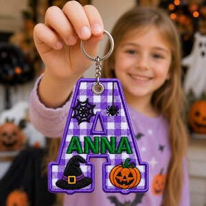 May include: A letter A shaped keyring featuring a purple and white gingham pattern. The letter is embroidered with the name ANNA in green letters and decorated with a black spider web, a small spider, a black witch hat, and an orange pumpkin.