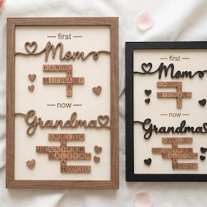 May include: Two framed wooden plaques with the words "Mom" and "Grandma" in cursive script. The plaques include names spelled out with Scrabble tiles and heart-shaped accents. One frame is brown, the other black.