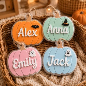 May include: Four pumpkin-shaped ornaments with white lettering. The orange pumpkin features a black bat, the mint green one has a black witch hat, the pink one displays a white ghost with a bow, and the light blue one shows a white ghost with a bowtie.