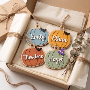 May include: Four wooden pumpkin-shaped ornaments with jute twine hangers, each featuring a name in white script: Emily on blue, Ethan on orange, Theodore on terracotta, and Hazel on sage green. Each pumpkin includes a small black bat or purple hat.