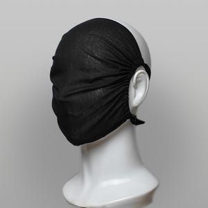 May include: A solid black fabric face mask covering the nose and mouth, featuring gathered side edges and thin fabric ties secured behind the ears. The mask is displayed on a smooth, white, featureless mannequin head against a neutral background.