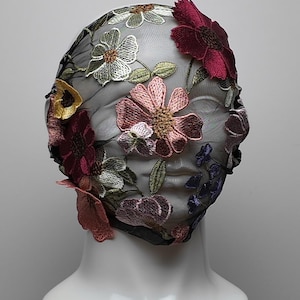 Può includere: Un copricapo in rete nera ornato di fiori ricamati nei toni del rosa, bordeaux, bianco e viola. Il design floreale è dettagliato con foglie verdi. Il copricapo è esposto su una testa di manichino bianca.