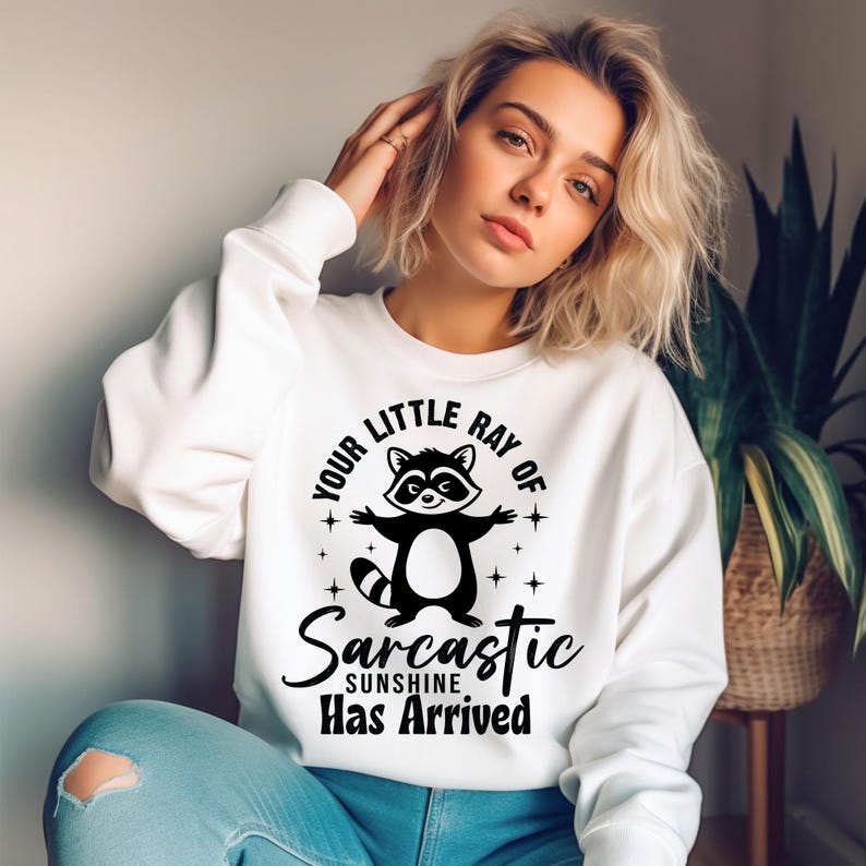May include: White sweatshirt with a black and white raccoon graphic and the text "Your Little Ray of Sarcastic Sunshine Has Arrived". The raccoon has its arms outstretched. The sweatshirt is a casual style.