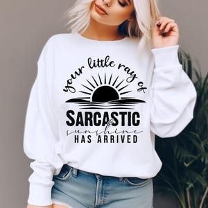May include: White sweatshirt with the text "your little ray of sarcastic sunshine has arrived" in black. The design features a sun graphic above the text. The sweatshirt is a casual style.