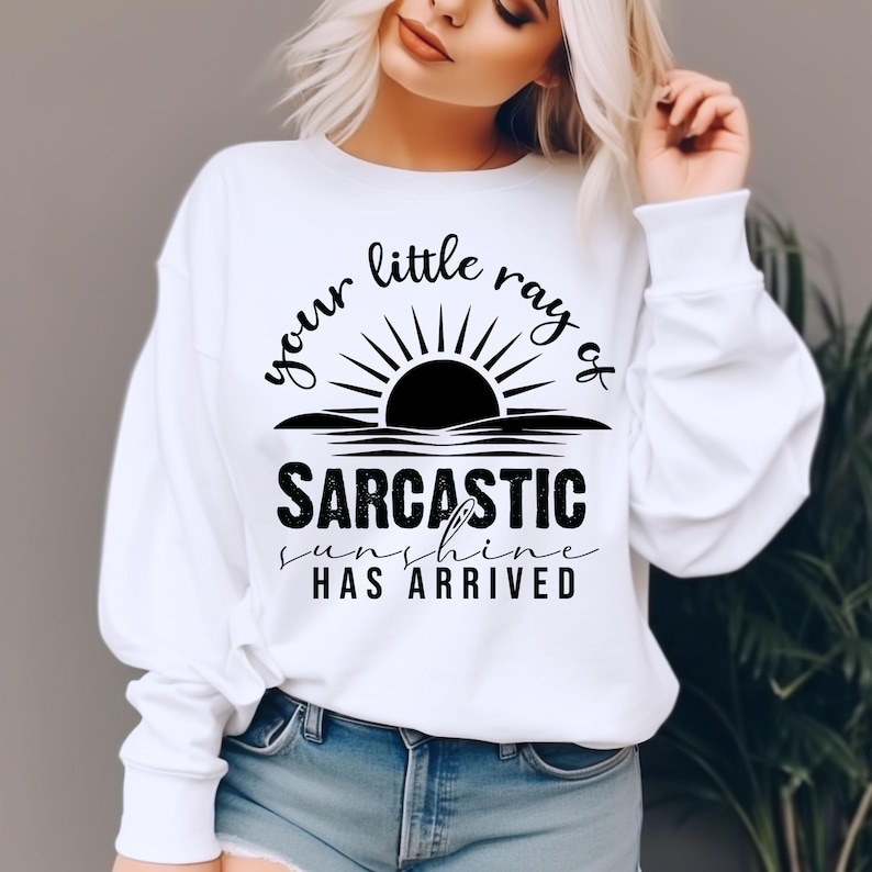 May include: White sweatshirt with the text "your little ray of sarcastic sunshine has arrived" in black. The design features a sun graphic above the text. The sweatshirt is a casual style.