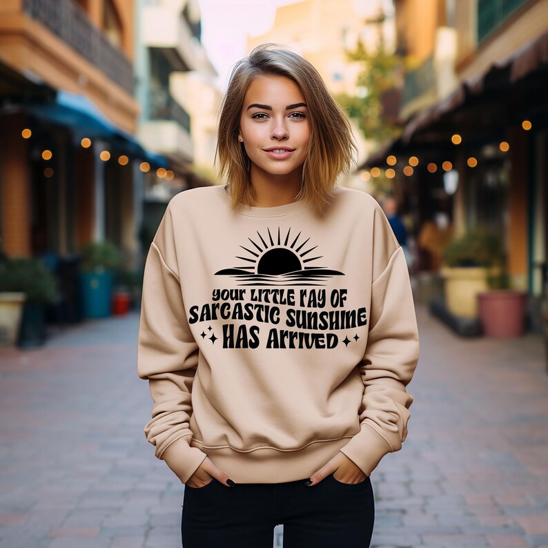 May include: A beige sweatshirt with the text "Your Little Ray of Sarcastic Sunshine Has Arrived" in black. The design includes a sun graphic. The sweatshirt is a casual style.