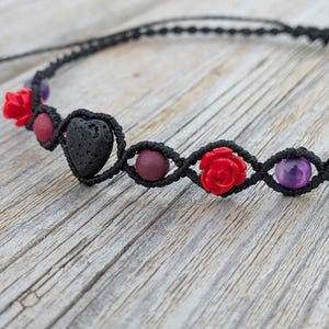 May include: A black macrame bracelet featuring a central black lava stone heart, interspersed with red rose-shaped beads, smooth dark red spherical beads, and polished purple spherical beads, all secured within a knotted black cord design.
