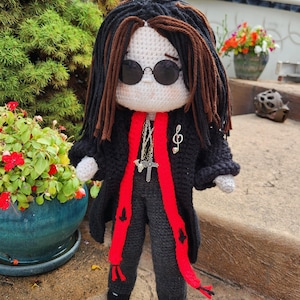 May include: Crocheted doll with black hair and sunglasses, wearing a black coat with a red scarf and gray pants. The doll has a cross necklace and a musical note pin. The doll is standing on a stone surface.