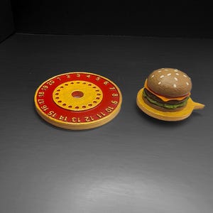 Puede incluir: Una pieza de juego redonda de madera roja y amarilla con n&uacute;meros del 1 al 20 alrededor del borde, y una peque&ntilde;a hamburguesa de madera sobre un plato de madera amarillo. La hamburguesa tiene pan de s&eacute;samo, queso y lechuga.