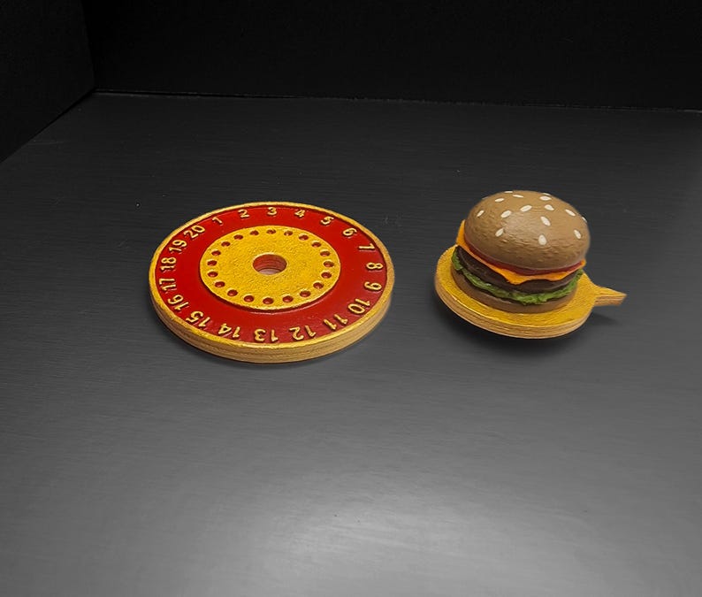 Puede incluir: Una pieza de juego redonda de madera roja y amarilla con n&uacute;meros del 1 al 20 alrededor del borde, y una peque&ntilde;a hamburguesa de madera sobre un plato de madera amarillo. La hamburguesa tiene pan de s&eacute;samo, queso y lechuga.