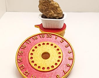 Puede incluir: Una esfera de reloj roja y dorada impresa en 3D con los números del 1 al 20. Un pequeño objeto dorado se encuentra encima de una plataforma roja, que a su vez está sobre la esfera del reloj. La esfera tiene un agujero central y un anillo de agujeros más pequeños.