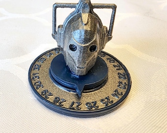 Puede incluir: Una figura de cabeza de Cyberman gris metálico sobre una base circular azul oscuro y dorada. La base presenta números alrededor del borde. La cabeza de Cyberman tiene una apariencia detallada y texturizada, con cuencas de los ojos y un marco rectangular.