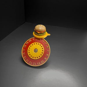 Puede incluir: Una hamburguesa en miniatura se encuentra sobre un objeto circular rojo y amarillo con los n&uacute;meros del 1 al 20. La hamburguesa tiene un pan con semillas de s&eacute;samo, una hamburguesa, queso y lechuga. El objeto tiene un agujero central y agujeros m&aacute;s peque&ntilde;os.