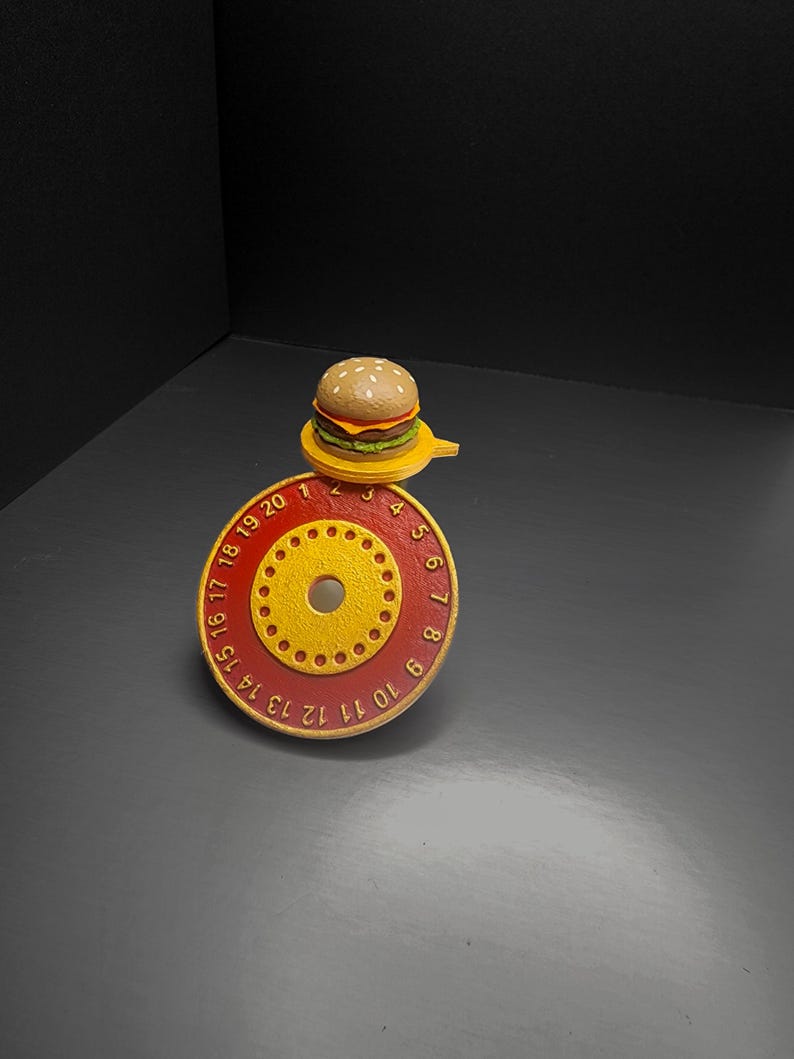 Puede incluir: Una hamburguesa en miniatura se encuentra sobre un objeto circular rojo y amarillo con los n&uacute;meros del 1 al 20. La hamburguesa tiene un pan con semillas de s&eacute;samo, una hamburguesa, queso y lechuga. El objeto tiene un agujero central y agujeros m&aacute;s peque&ntilde;os.