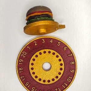 Puede incluir: Un objeto dorado en forma de hamburguesa con una base roja y dorada. La base tiene los n&uacute;meros 1-20 alrededor del borde. La hamburguesa tiene pan, carne y aderezos. El objeto es probablemente un temporizador de cocina o un art&iacute;culo decorativo.
