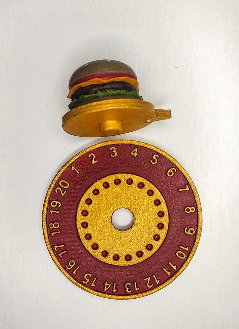 Puede incluir: Un objeto dorado en forma de hamburguesa con una base roja y dorada. La base tiene los n&uacute;meros 1-20 alrededor del borde. La hamburguesa tiene pan, carne y aderezos. El objeto es probablemente un temporizador de cocina o un art&iacute;culo decorativo.