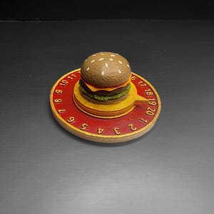 Puede incluir: Un reloj novedoso con forma de hamburguesa con una base roja y dorada. La esfera del reloj presenta n&uacute;meros y una peque&ntilde;a hamburguesa en la parte superior. La hamburguesa tiene un pan de semillas de s&eacute;samo, una hamburguesa, queso y lechuga.