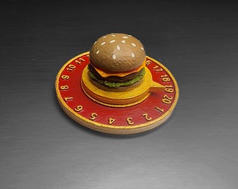 May include: A novelty burger-shaped clock with a red and gold base. The clock face features numbers and a small burger on top. The burger has a sesame seed bun, a patty, cheese, and lettuce. The clock's hands are not visible.