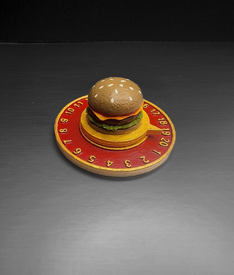 Puede incluir: Un reloj novedoso con forma de hamburguesa con una base roja y dorada. La esfera del reloj presenta n&uacute;meros y una peque&ntilde;a hamburguesa en la parte superior. La hamburguesa tiene un pan de semillas de s&eacute;samo, una hamburguesa, queso y lechuga.