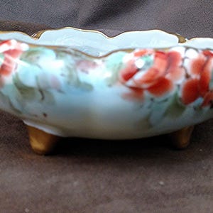 May include: A small, round, footed porcelain bowl featuring a light blue base color with hand-painted red and pink floral motifs. The bowl has a scalloped rim with gold trim and three short, gold-painted feet supporting the base.