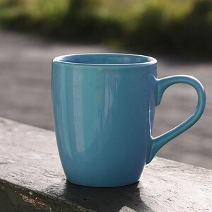 May include: A solid turquoise ceramic mug with a curved handle. The mug has a glossy finish and is sitting on a weathered surface. The mug is designed for hot beverages and is suitable for home or office use.