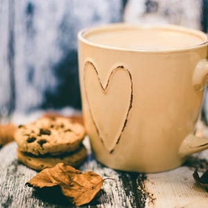 May include: A beige ceramic mug with a heart-shaped design, filled with a creamy beverage. Two chocolate chip cookies and dried leaves are on a rustic wooden surface. The mug has a large handle.