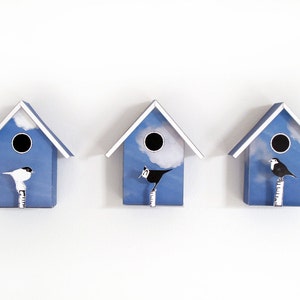 May include: Three blue birdhouses with white trim, each with a different bird perched on a branch. The birdhouses have a sky blue background with white clouds and a round opening.