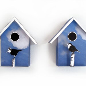 May include: Two blue birdhouses with white trim and a white cloud design. Each birdhouse has a black bird perched on a white pole.