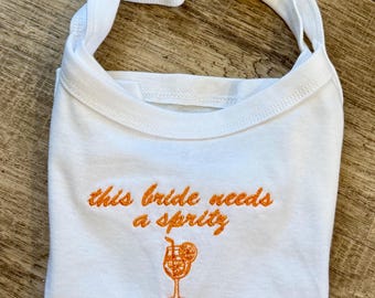 May include: A white tank top featuring orange embroidery that reads this bride needs a spritz, accompanied by an embroidered orange cocktail glass graphic with a straw and a citrus slice garnish, centered on the front of the garment.