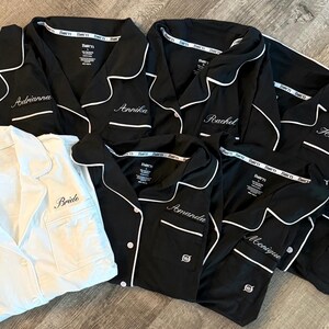 May include: A collection of black pajama sets with white piping and personalized names embroidered on the chest pockets. One white pajama set with "Bride" embroidered on the pocket is also included. The pajamas are laid out on a wooden surface.