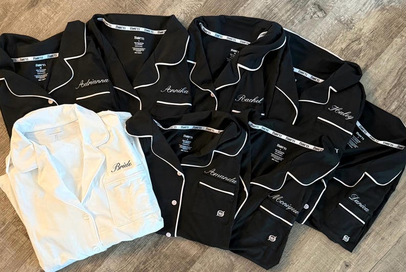 May include: A collection of black pajama sets with white piping and personalized names embroidered on the chest pockets. One white pajama set with "Bride" embroidered on the pocket is also included. The pajamas are laid out on a wooden surface.