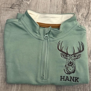 May include: A sage green zip-up sweatshirt with a white collar and brown trim. The sweatshirt features an embroidered deer head design and the name "HANK" in black lettering. The zipper is centered, and the garment is folded.