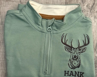May include: A sage green zip-up sweatshirt with a white collar and brown trim. The sweatshirt features an embroidered deer head design and the name "HANK" in black lettering. The zipper is centered, and the garment is folded.
