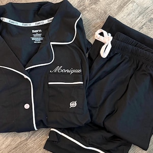 May include: Black pajama set with white piping and trim. The top has a breast pocket and the name "Monique" embroidered on the front. The pajama pants have a drawstring waist. The brand name "Born" is visible on the top.