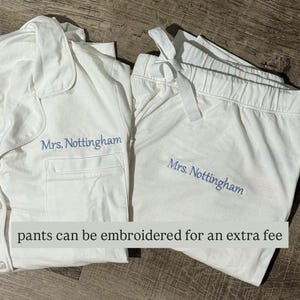 May include: White pajama set with a button-down top and drawstring pants. The text "Mrs. Nottingham" is embroidered in light blue on both pieces. The text "pants can be embroidered for an extra fee" is also visible.