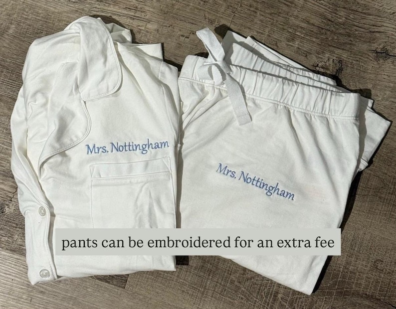 May include: White pajama set with a button-down top and drawstring pants. The text "Mrs. Nottingham" is embroidered in light blue on both pieces. The text "pants can be embroidered for an extra fee" is also visible.