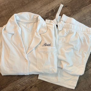 May include: White pajama set with a collared button-down shirt and matching pants. The shirt has a breast pocket and the word "Bride" embroidered in black script. The set is made of a soft, lightweight fabric.