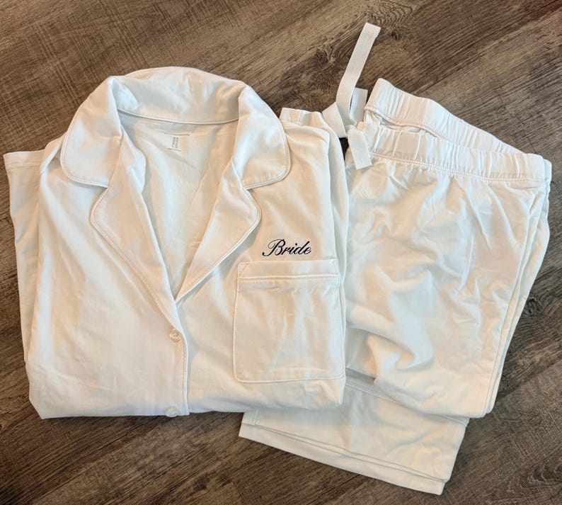 May include: White pajama set with a collared button-down shirt and matching pants. The shirt has a breast pocket and the word "Bride" embroidered in black script. The set is made of a soft, lightweight fabric.