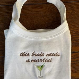 May include: White bib with the embroidered text "this bride needs a martini" in brown thread. A green martini glass with a lime and olive is embroidered below the text. The bib has a rounded neckline and two straps.