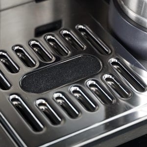 Könnte beinhalten: Nahaufnahme einer Abtropfschale aus Edelstahl mit einer Reihe länglicher Öffnungen und einem strukturierten schwarzen Mittelteil. Die Schale ist Teil eines größeren Geräts, wobei ein Teil einer metallischen Komponente im Hintergrund sichtbar ist.