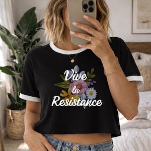 Puede incluir: Top corto negro con ribete blanco y la frase "Vive la Resistance" en escritura blanca con un diseño floral. La camiseta tiene mangas cortas y cuello redondo.