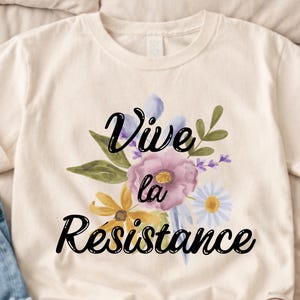 Puede incluir: Camiseta color crema con un diseño floral con flores moradas, amarillas y blancas. Las palabras "Vive la Résistance" están escritas en negro sobre las flores. La camiseta está sobre una superficie clara.